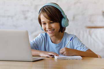 Happy adorable teenage kid boy in wireless headphones looking at laptop screen, listening educational online lecture, enjoying studying distantly at home, writing notes in copybook, e-learning concept © fizkes