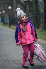 Little girl with a backpack, in a jacket and a hat near the school.