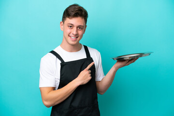 Young waitress with tray isolated on blue background pointing to the side to present a product