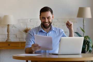 Emotional laughing happy young man looking at paper document, feeling excited reading amazing news,...