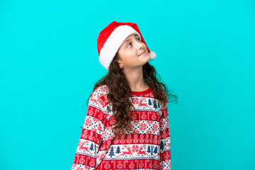 Little caucasian with christmas hat isolated on blue background looking up while smiling
