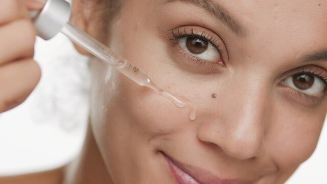 Cute slim young African American woman drips skin care serum on her cheek from dropper smiling for the camera on white background | Skin care serum commercial