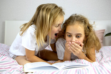 two cute little sisters with blond hair lie on the bed and read a book, tell scary stories and...