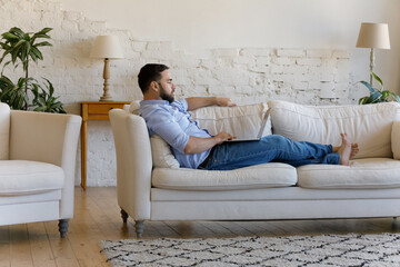 Fototapeta premium Full length concentrated young man holding computer on laps lying on cozy couch at home, enjoying working distantly on online project, communicating in social network or shopping in internet store.