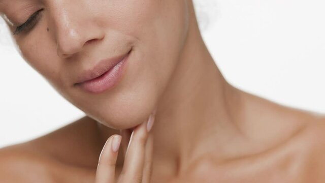 Horizontal big close-up of attractive slim young black-haired African American woman strokes her jaw line and turns her head to the camera smiling on white background | Sagging face prevention concept