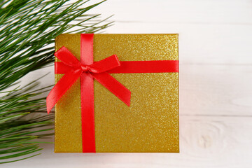 A beautiful golden gift box tied with a red bow or ribbon for holidays on a white wooden background and branches of a Christmas tree. Christmas Gift Concept