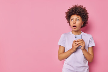 Horizontal shot of scared stunned woman holds mobile phone sees something scary wears casual t...