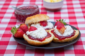 Scones with strawberry jam, cream and fresh strawberries on a red and white checkered tablecloth