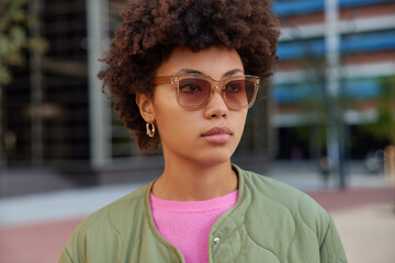 Serious curly haired young woman wears sunglasses and jacket has promenade in urban setting...