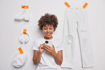 Curious curly haired woman looks with interest at smartphone screen bites lips reads message content dressed in casual t shirt poses against white background with items of clothes plastered.