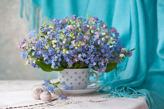 A bouquet of spring flowers forget-me-nots and lilies of the valley in a mug on a table with a tablecloth against the background of a wall and a shawl. Beautiful card.