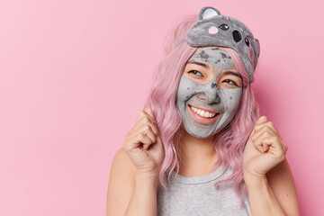 Indoor shot of happy woman clenches fists celebrates good news applies clay mask for skin rejuvenation has joyful dreamy expression wears sleepmask on forehead poses against pink studio wall
