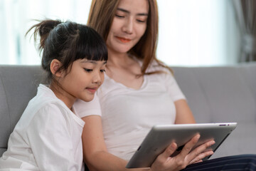 Fototapeta premium Beautiful Asian mother with daughter sitting on the sofa and playing video game on a tablet while spending time together at home