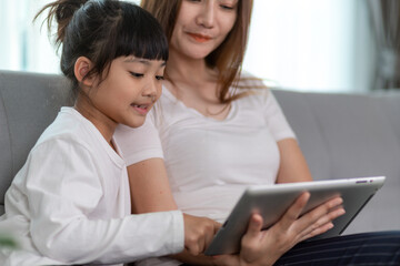 Beautiful Asian mother with daughter sitting on the sofa and playing video game on a tablet while spending time together at home