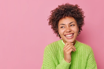 Portrait of happy woman with curly hair holds chin smiles broadly with teeth feels sincere dressed in knitted green sweater poses against pink background blank copy space for your promotional content