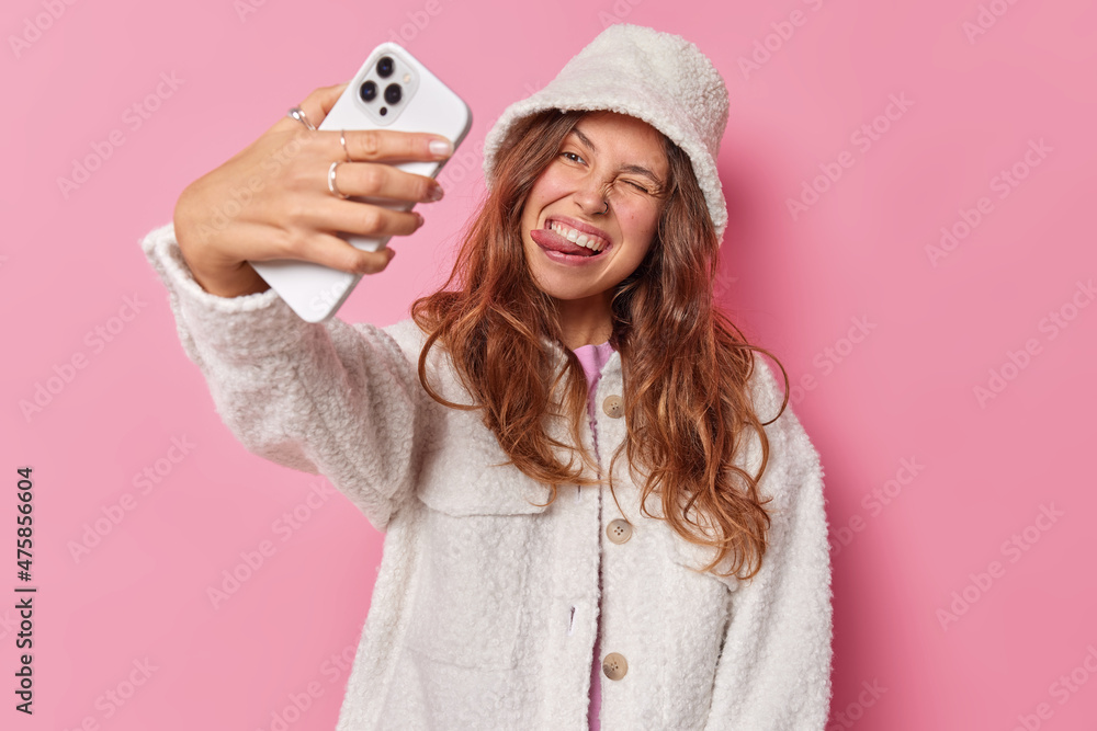 Wall mural optimistic carefree woman sticks out tongue winks eye poses at smartphone camera makes selfie portra