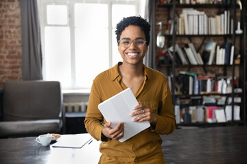 Portrait of joyful young African American woman in eyeglasses holding digital computer tablet in...