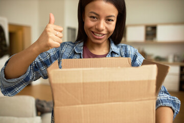Happy cute mixed-race woman showing thumb up holding unpacked carton parcel, satisfied and pleased...