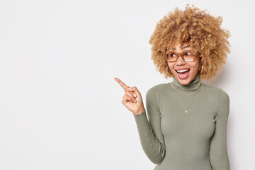 Happy surprised female promoter points away at blank copy space shows something awesome demonstrates mock up place wears eyewear and turtleneck isolated over white background shows promo sale