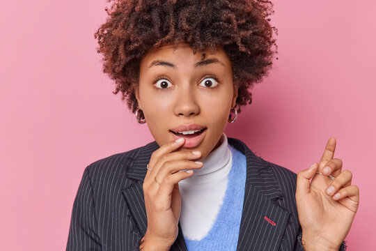 Portrait of surprised curly haired woman looks with widely opened eyes feels worried stares bugge eyes dressed elegantly isolated over pink background gasps fascinated. Human reactions concept