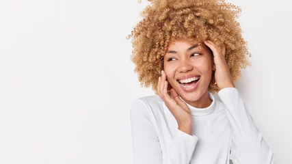 Fotobehang Positive European woman with curly hair touches face gently looks happily away wears casual jumper feels glad has carefree glad expression poses against white background with copy space area © Wayhome Studio