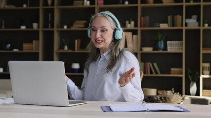 Positive retired female in headset hold distant english class to remote students explain learning material speaking before laptop webcam. Smiling aged woman teach pupils on distance via web conference