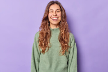 Positive young woman with long wavy hair dressed in casual jumper feels very happy being in good mood hears compliment isolated over purple background watches something funny. Emotions concept