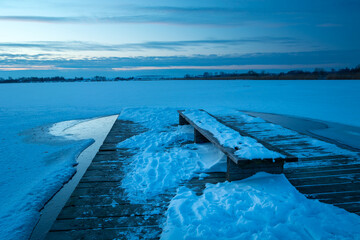 Snow on the pier with a bench and a frozen lake