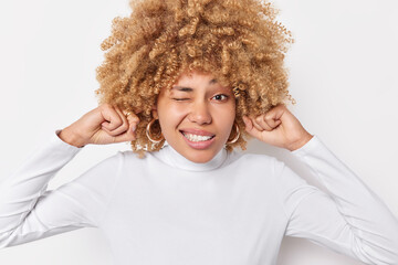 Displeased curly haired blonde woman plugs ears avoids noisy sound avoids loud noise has irriatated expression dressed in casual jumper isolated over white background. Body language concept.