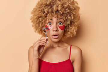 Stunned emotional curly woman going to do makeup uses eyelashes curler applies hydrogel patches to reduce wrinkles dressed in red t shirt poses against beige background. Beauty routines concept