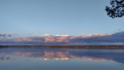 Fototapeta premium There are ripples on the water of the lake. Evening is coming. On the far shore the forest and over it the moon has risen and clouds have accumulated. A pine branch is leaning over the water