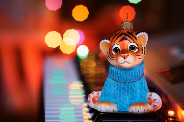 An orange tiger cub in a blue sweater sits on a musical synthesizer. In the background the lights of an electric Christmas garland. Reflection of light of lights. Greeting card. Selective focus.