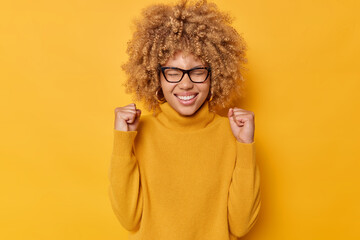 Fototapeta Waist up shot of euphoric positive young woman clenches fists celebrates achievements reaches goal exclaims yes smiles broadly wears eyeglasses casual jumper isolated over yellow background. obraz
