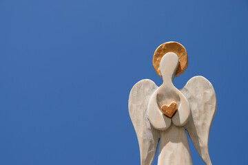 Wooden angel with golden halo and heart in hands. Blue sky background.