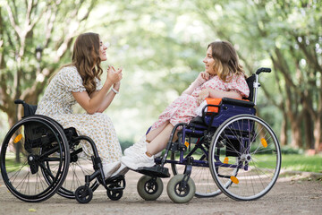 Side view of two happy women in wheelchairs talking and smiling outdoors. Background of green...