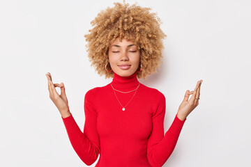 Calm concentrated woman with curly hair keeps eyes closed and meditates indoor makes mudra gesture...