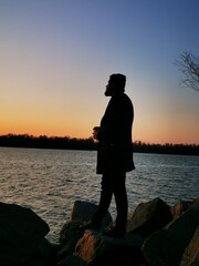 A man stands on the beach with coffee admiring the wonderful sunset over the lake and meditates. A wonderful trip to nature. Back view, backlight