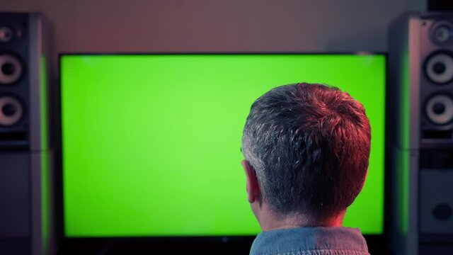 Rear view of the head of a man sitting facing a large blank green screen of a comouter or television in a conceptual image