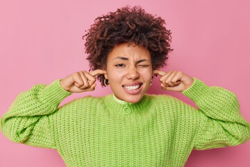 Displeased curly haired young woman plugs ears cannot focus in crowded place feels discomfort over...