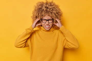 Photo of displeased woman keeps hands on temples suffers from migraine clenches teeth to reveal pain cannot concentrate in noisy atmosphere dressed in casual jumper isolated over yellow wall