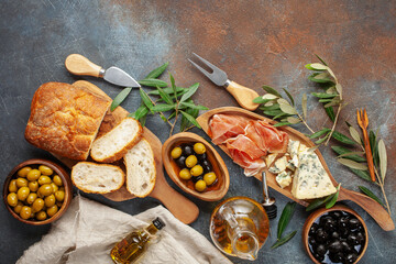 Black and green Olives, olive tree branch, oil, cheese Dorblu or Gorgonzola, prosciutto and ciabatta bread on dark  table. Top view. Flat lay