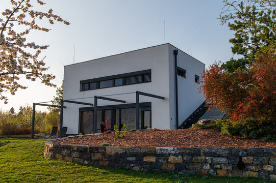 Modern, white, cube, elegant, minimalist style passive house with large panoramic windows, grey shutters in maintained garden in sunset warm light. Wooden terrace. Garden furniture.