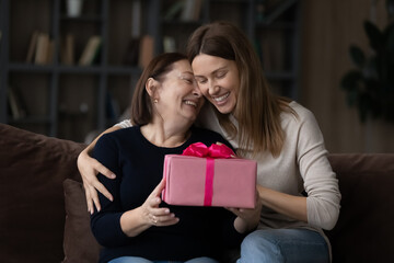 Happy birthday mommy. Affectionate young woman adult daughter sit on sofa embrace excited older mum greet aged parent with Christmas. Caring female grown child give senior mom present box on March 8 - obrazy, fototapety, plakaty