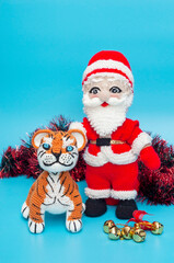 On a blue background there are knitted toys of Santa Claus and a tiger next to tinsel and a Christmas tree toy. vertical snapshot.