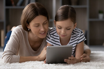 Safe internet for kids. Interested single mom nanny grown sister hug preteen girl lie on floor watch video online scroll social media on pad together. Small girl use tablet pc under control of mum