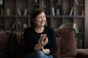 Pleasant dreams. Happy mature female retiree sit on sofa hold modern mobile phone gadget smile look at distance plan to make call to adult child. Thoughtful old lady with cell lost in good thoughts