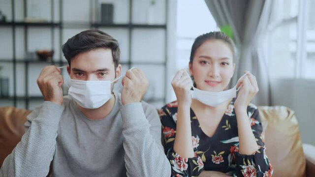 Hand talking face mask protection off,asian and caucasian couple sitting on sofa look at camera hand pull protective face mask off with happiness and cheerful after covid spread is over lock down over