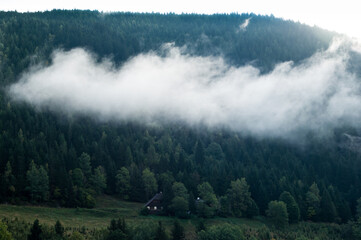 Panoramablick auf eine bergige Landschaft , mit Nadelbäumen und Wiesen, mit einem einsam stehenden Haus und sehr tief hängenden Wolken