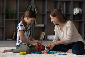 Good weekend together. Happy loving young mom and tween child daughter play exciting brain developing game on carpet at home. Grown elder and younger sisters engaged in building from toy constructor