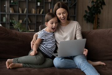 Wow. Excited single mother and tween daughter sit on couch read amazing news on laptop screen win lottery get prize from web store. Surprised shocked nanny and little girl play online videogame on pc